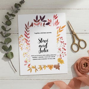 Puede incluir: Una invitación de boda con un diseño floral en colores otoñales. La invitación presenta los nombres "Steve y Julie" y los detalles del evento. Tijeras doradas y una cinta sobre una superficie de madera blanca.