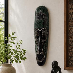 Peut inclure: Un masque mural en bois long et étroit, de couleur vert foncé et noir, orné de motifs pointillés complexes et d'un oiseau stylisé. Le masque présente une finition lisse et polie avec des traits du visage marqués, dont des yeux en amande.