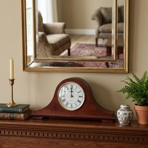 May include: A mahogany-colored wooden mantel clock with a rounded top, a white circular face, black Roman numerals, and black hands. The clock is positioned on a wooden surface alongside a brass candlestick, two books, a small patterned jar, and a fern.