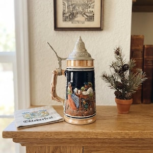Op de afbeelding: Een keramische bierpul met een donkerblauwe behuizing voorzien van een reliëfscène van mensen aan een tafel. De pul heeft een metalen conisch deksel en een handvat in de vorm van een boomtak. Daarnaast ligt een stoffen servet met de tekst Oktoberfest.
