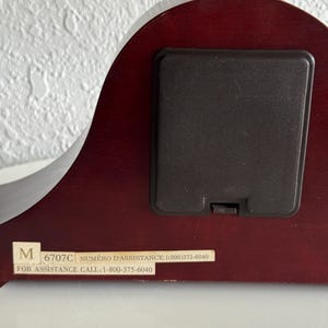 May include: The back of a dark cherry-toned wooden clock featuring a rectangular black battery compartment cover. A white rectangular label is affixed to the bottom, displaying the text M 6707C NUM&Eacute;RO D'ASSISTANCE: 1(800)375-6040 FOR ASSISTANCE CALL: 1-800-375-6040.