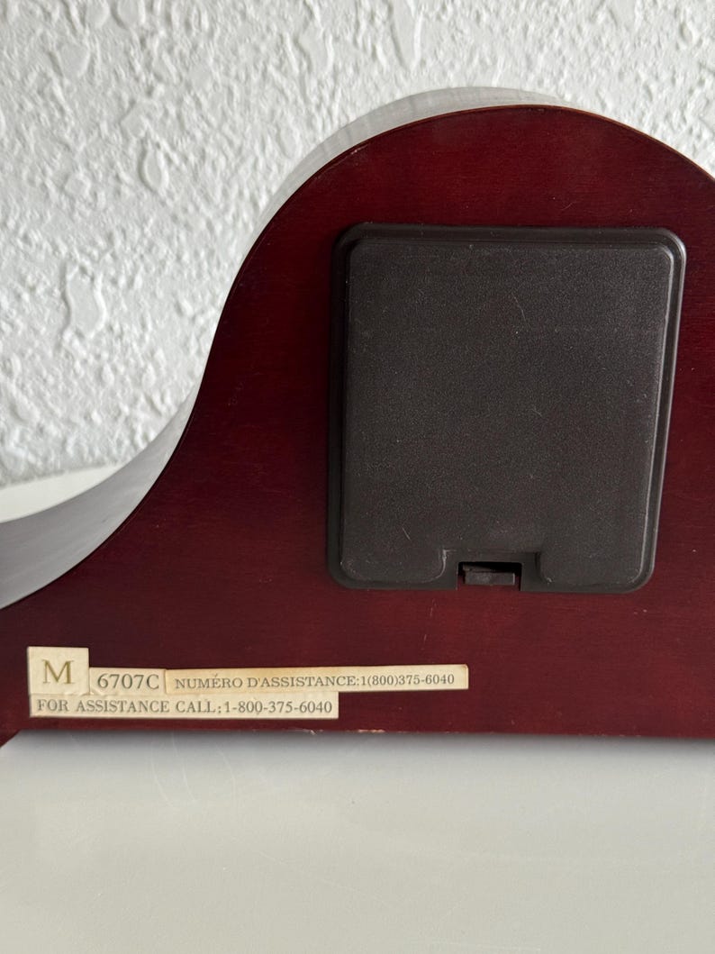 May include: The back of a dark cherry-toned wooden clock featuring a rectangular black battery compartment cover. A white rectangular label is affixed to the bottom, displaying the text M 6707C NUM&Eacute;RO D'ASSISTANCE: 1(800)375-6040 FOR ASSISTANCE CALL: 1-800-375-6040.