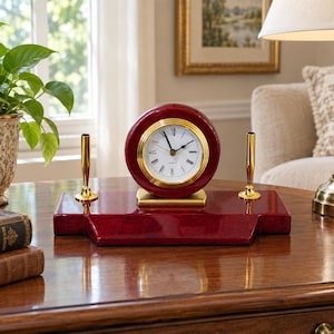 May include: A desk clock set featuring a circular, deep red clock face with a gold-toned bezel and Roman numerals, mounted on a matching rectangular red base. Two gold-toned pen holders are positioned on either side of the clock on the base.