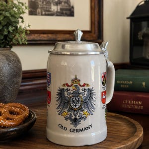 以下が含まれることがあります： 金属製の蓋と取っ手が付いたライトグレーの陶器製ビールジョッキ。正面には詳細な鷲の紋章があり、その下に「OLD GERMANY」という文字が印刷されています。ジョッキの側面には、より小さな地域の紋章が見えます。