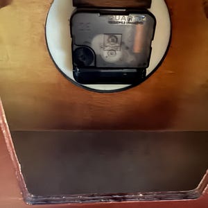 May include: A dark brown wooden clock housing with a black plastic quartz movement mechanism mounted in a circular cutout. The back panel features a rectangular opening and a white sticker with the text SISTANCE:1(800)375-6040 at the bottom.