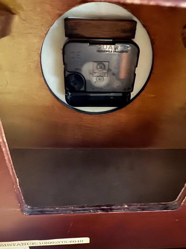 May include: A dark brown wooden clock housing with a black plastic quartz movement mechanism mounted in a circular cutout. The back panel features a rectangular opening and a white sticker with the text SISTANCE:1(800)375-6040 at the bottom.