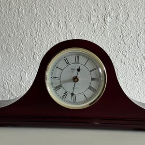 May include: A dark wood mantel clock with an arched top and a circular white face. The clock features black Roman numerals, thin black hands, and a gold-toned bezel. The text "Quartz" and a brand name are printed on the center of the clock face.