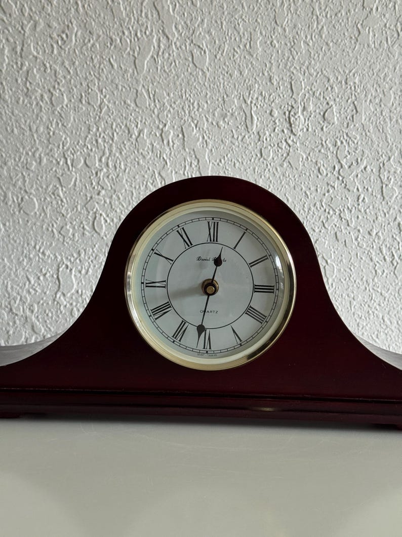 May include: A dark wood mantel clock with an arched top and a circular white face. The clock features black Roman numerals, thin black hands, and a gold-toned bezel. The text "Quartz" and a brand name are printed on the center of the clock face.