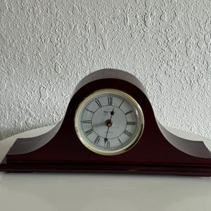 May include: A dark brown wooden mantel clock with a curved top and a rectangular base. The clock face is white with black Roman numerals, a gold-toned circular bezel, and black hands. The text "Rhythm Quartz" is printed on the clock face.