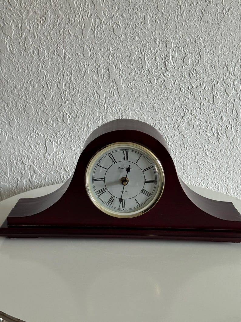 May include: A dark brown wooden mantel clock with a curved top and a rectangular base. The clock face is white with black Roman numerals, a gold-toned circular bezel, and black hands. The text "Rhythm Quartz" is printed on the clock face.