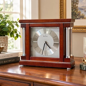 May include: A rectangular mantel clock with a dark cherry wood frame and silver-toned metal accents. The clock face features a circular silver dial with thin black hour markers and black hands. The word Bulova is printed in black on the dial.