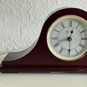 May include: A dark mahogany-colored wooden mantel clock with a curved top. The clock face is white with black Roman numerals, gold-colored hands, and the text Daniel Richards and Quartz printed in black. It features a gold-toned circular bezel.