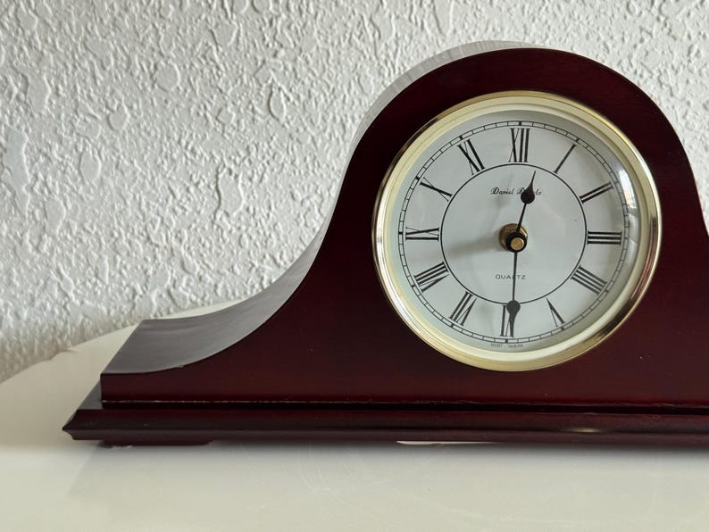 May include: A dark mahogany-colored wooden mantel clock with a curved top. The clock face is white with black Roman numerals, gold-colored hands, and the text Daniel Richards and Quartz printed in black. It features a gold-toned circular bezel.