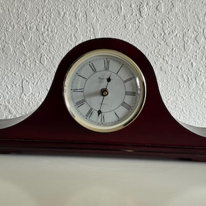 May include: A dark mahogany-colored wooden mantel clock with an arched top. The clock face is white with black Roman numerals, a gold-toned bezel, and black hour and minute hands. The text Royal Quartz is printed in black on the clock face.