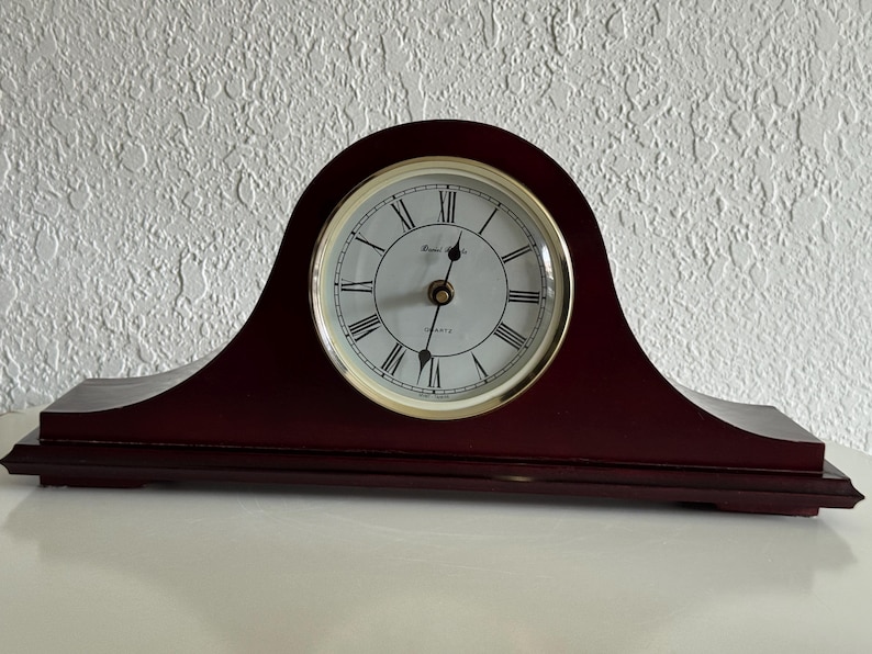 May include: A dark mahogany-colored wooden mantel clock with an arched top. The clock face is white with black Roman numerals, a gold-toned bezel, and black hour and minute hands. The text Royal Quartz is printed in black on the clock face.