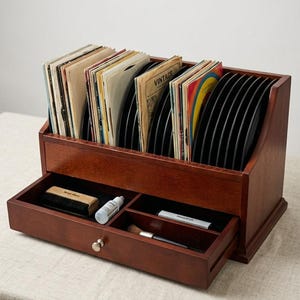 Peut inclure: Un support de rangement pour disques vinyles en bois foncé avec une section supérieure pour plusieurs disques et un tiroir contenant une brosse de nettoyage, un petit flacon de liquide nettoyant et une brosse pour stylet. Le mot VINTAGE est inscrit.