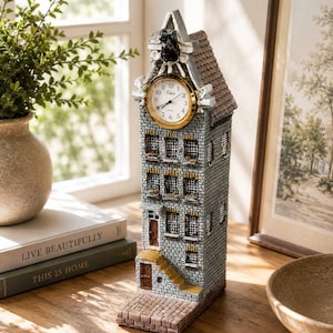 Pode incluir: Um relógio decorativo em forma de um edifício estreito de tijolo, com um mostrador dourado e branco. O edifício tem um telhado castanho e pequenas janelas. O relógio está numa superfície de madeira.