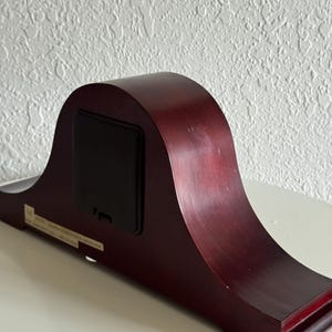 May include: A dark cherry-toned wooden mantel clock viewed from the back, featuring a smooth, arched silhouette and a rectangular black battery compartment cover centered on the rear panel, with a small white informational sticker attached below.