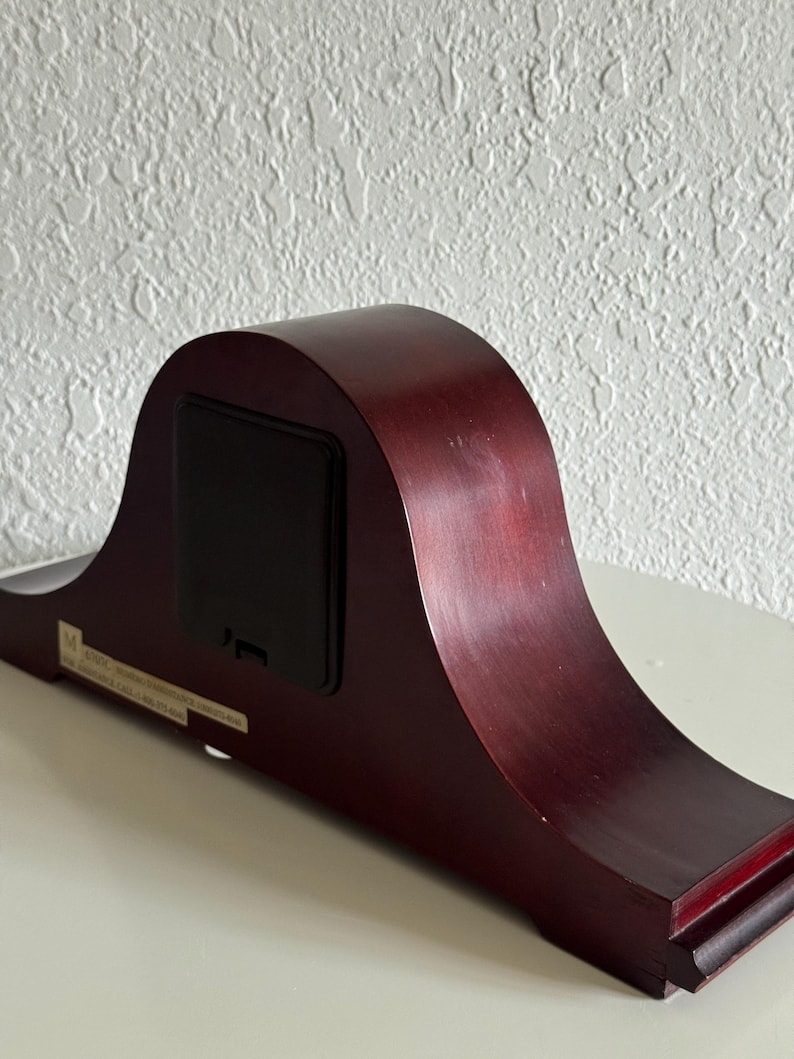 May include: A dark cherry-toned wooden mantel clock viewed from the back, featuring a smooth, arched silhouette and a rectangular black battery compartment cover centered on the rear panel, with a small white informational sticker attached below.
