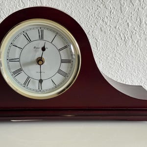May include: A dark cherry-colored wooden mantel clock with a curved top. The clock face is white with black Roman numerals, gold-colored hands, and a gold-toned bezel. The text Daniel Steels and QUARTZ are printed on the white clock face.