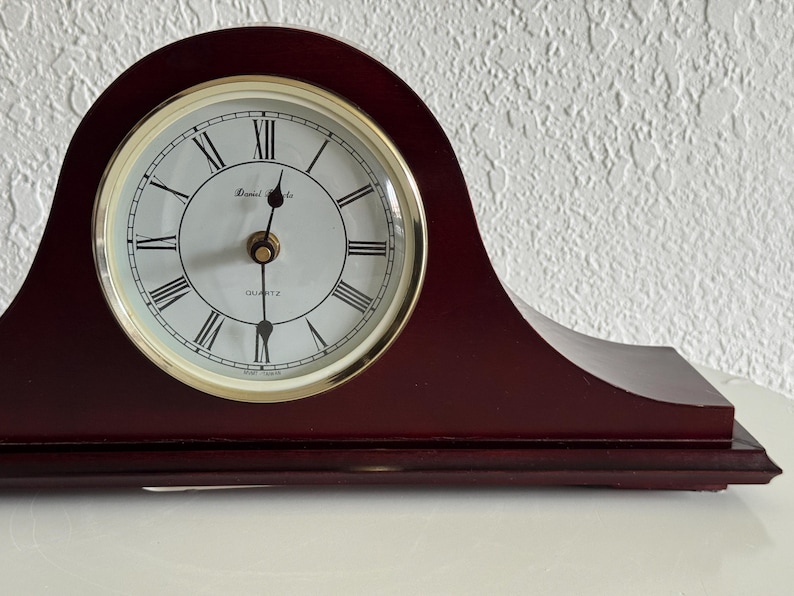 May include: A dark cherry-colored wooden mantel clock with a curved top. The clock face is white with black Roman numerals, gold-colored hands, and a gold-toned bezel. The text Daniel Steels and QUARTZ are printed on the white clock face.