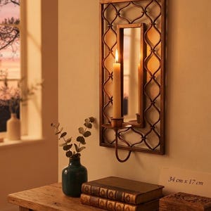 May include: A rectangular wall-mounted candle holder with a bronze-toned metal frame featuring an intricate lattice pattern and a central mirror. A single lit taper candle sits in the attached cup. The dimensions are 34 cm x 17 cm.