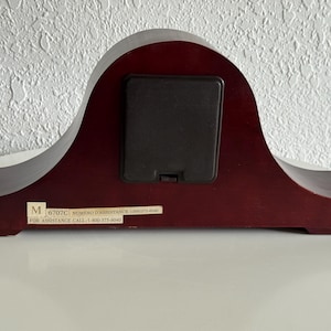 May include: A dark mahogany-colored wooden mantel clock back featuring a central black square battery compartment cover. A rectangular white sticker at the bottom displays the letter M, the code 6707C, and the text FOR ASSISTANCE CALL 1-800-375-6040.