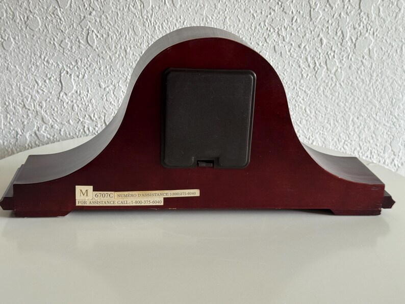 May include: A dark mahogany-colored wooden mantel clock back featuring a central black square battery compartment cover. A rectangular white sticker at the bottom displays the letter M, the code 6707C, and the text FOR ASSISTANCE CALL 1-800-375-6040.