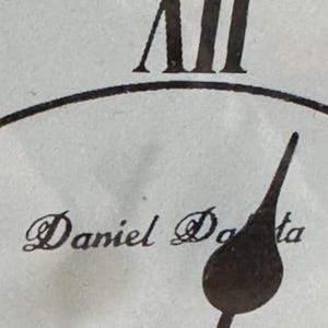May include: A close-up of a clock face featuring the Roman numeral XII at the top, a dark metal clock hand pointing downwards, and the text Daniel Doda printed in a black serif font on a light, textured off-white background surface.
