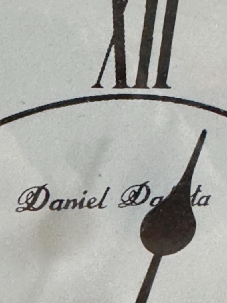 May include: A close-up of a clock face featuring the Roman numeral XII at the top, a dark metal clock hand pointing downwards, and the text Daniel Doda printed in a black serif font on a light, textured off-white background surface.