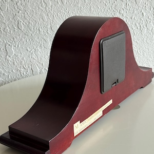 May include: A dark wood mantel clock viewed from the back, featuring a smooth arched silhouette, a rectangular black battery compartment cover, and a white rectangular label with the text M 6707C NUMERO D'ASSISTANCE 1-800-575-8040.