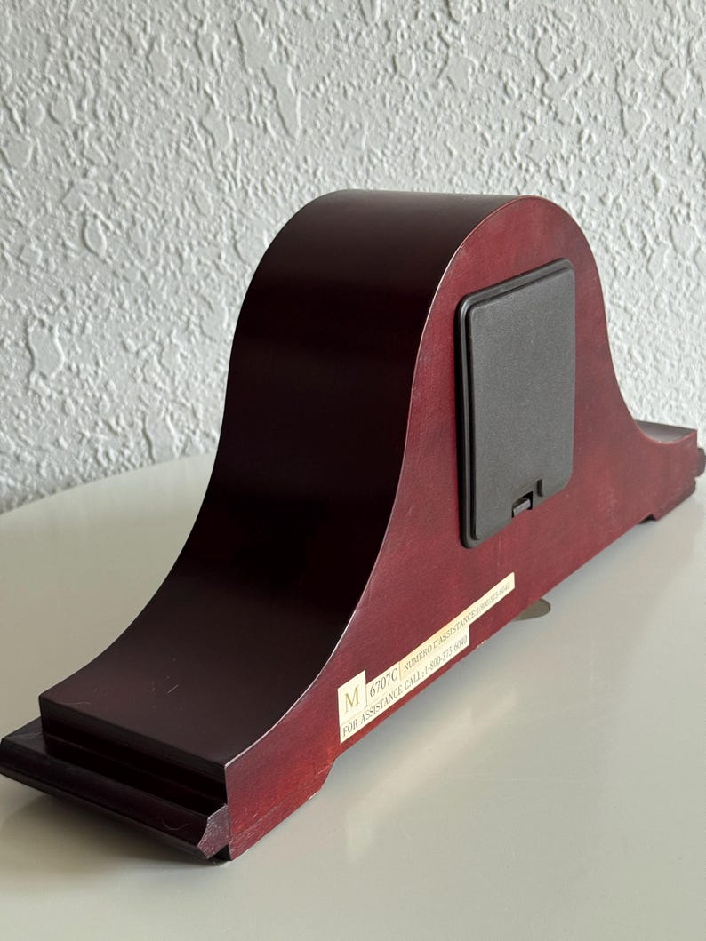 May include: A dark wood mantel clock viewed from the back, featuring a smooth arched silhouette, a rectangular black battery compartment cover, and a white rectangular label with the text M 6707C NUMERO D'ASSISTANCE 1-800-575-8040.
