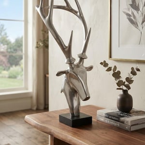 May include: A silver-toned deer head sculpture with prominent antlers, mounted on a solid black rectangular base. The metallic finish is smooth and reflective, capturing the light across the curved surfaces of the neck and the branching antlers.