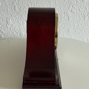 May include: A side view of a dark brown wooden mantel clock with a smooth, arched top and a rectangular base. The clock features a circular gold-toned metal bezel on the side, showcasing a polished finish that contrasts with the deep wood grain.