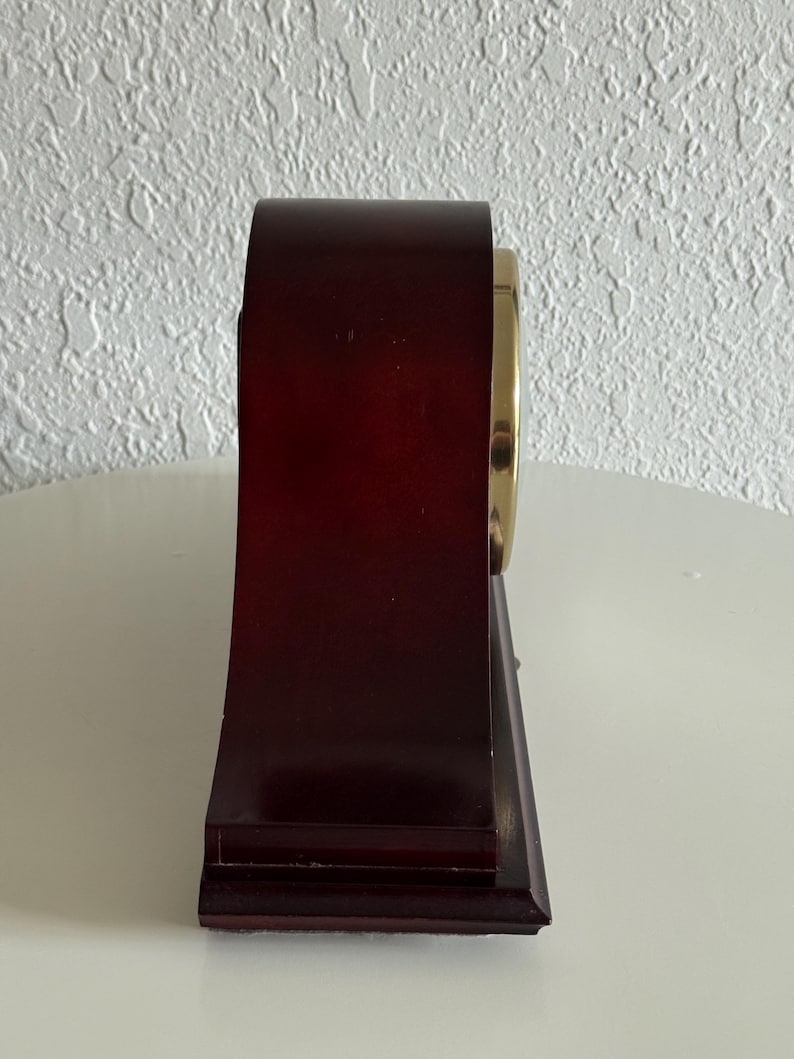 May include: A side view of a dark brown wooden mantel clock with a smooth, arched top and a rectangular base. The clock features a circular gold-toned metal bezel on the side, showcasing a polished finish that contrasts with the deep wood grain.