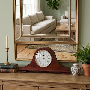 May include: A dark wood mantel clock with an arched top and a white circular face featuring black Roman numerals and gold hands. The clock is positioned on a wooden surface alongside two stacked books, a brass candle holder, and a small plant.