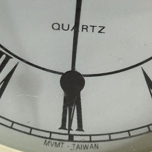 May include: A close-up of a white clock face featuring black Roman numerals, black hands, and the text QUARTZ printed in black above the center. The text MVMT TAIWAN is printed in black at the bottom edge of the dial, near the six o'clock position.