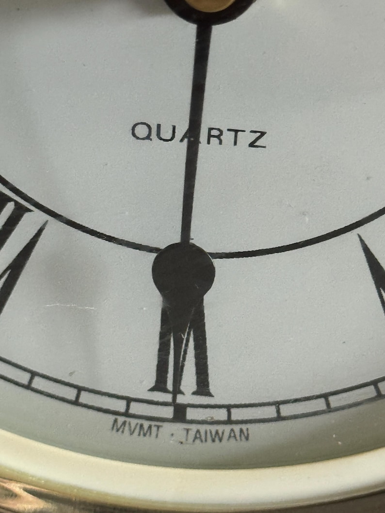 May include: A close-up of a white clock face featuring black Roman numerals, black hands, and the text QUARTZ printed in black above the center. The text MVMT TAIWAN is printed in black at the bottom edge of the dial, near the six o'clock position.