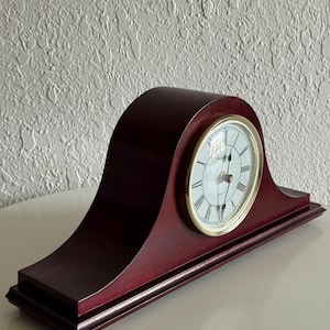 May include: A dark cherry-colored wooden mantel clock with a curved top and a rectangular base. The clock face is white with black Roman numerals, black hands, and a gold-toned circular bezel. The word Quartz is printed on the clock face.