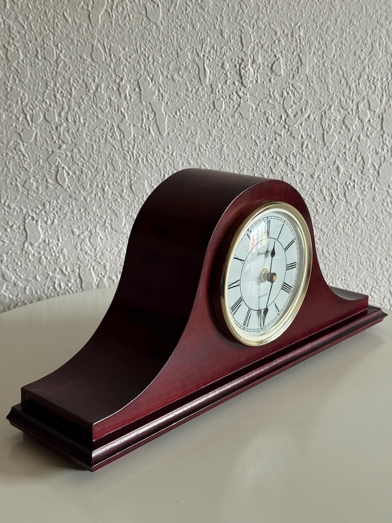 May include: A dark cherry-colored wooden mantel clock with a curved top and a rectangular base. The clock face is white with black Roman numerals, black hands, and a gold-toned circular bezel. The word Quartz is printed on the clock face.