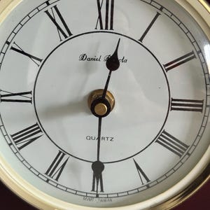 May include: A round analog clock face with a white background and black Roman numerals. The clock features black hour and minute hands, a gold-colored center cap, and the text Daniel Dakota, QUARTZ, and MVMT TAIWAN printed on the dial.