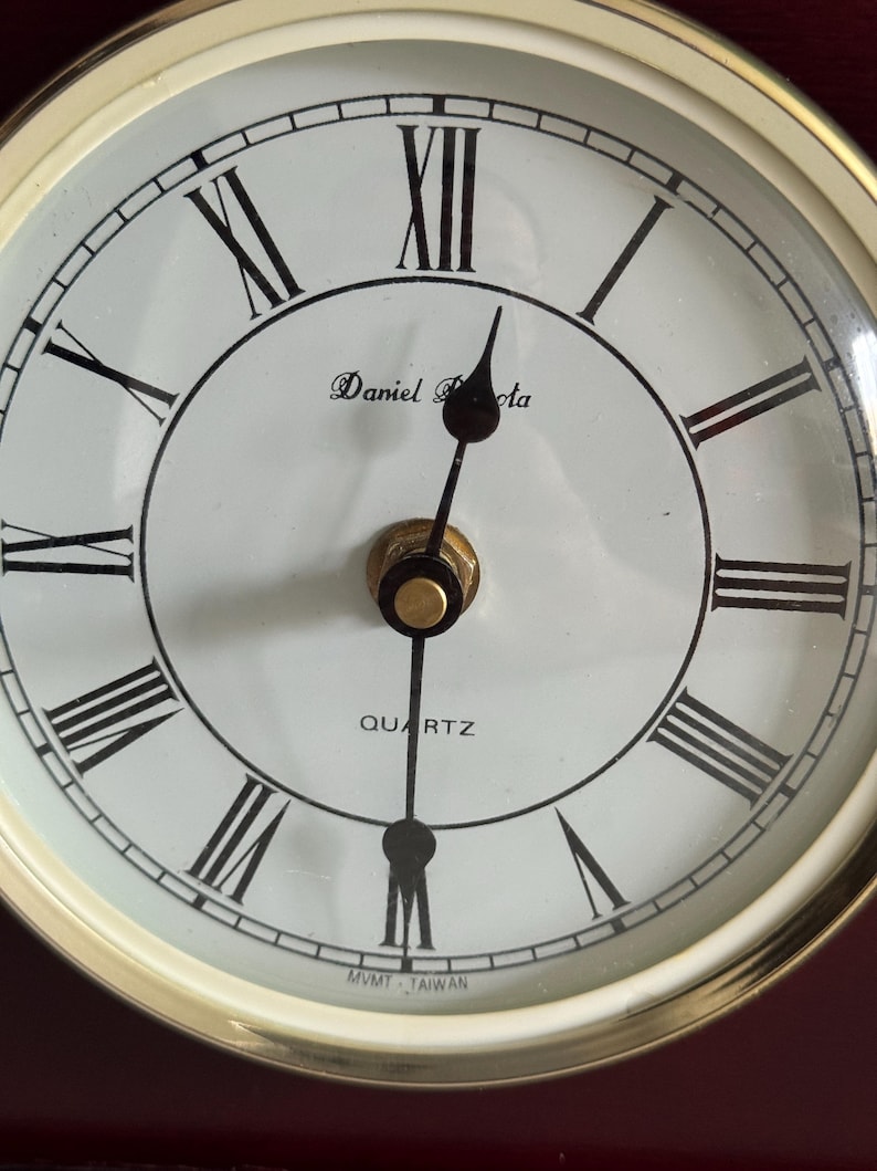 May include: A round analog clock face with a white background and black Roman numerals. The clock features black hour and minute hands, a gold-colored center cap, and the text Daniel Dakota, QUARTZ, and MVMT TAIWAN printed on the dial.