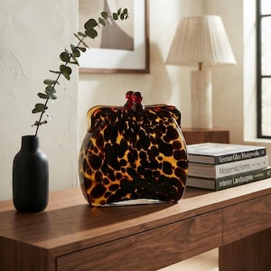 Puede incluir: Un objeto decorativo de vidrio con forma de monedero vintage con un patrón de carey marrón y negro. La pieza presenta una silueta redondeada y dos pequeños pomos esféricos de color rojo oscuro en la parte superior, imitando un cierre.
