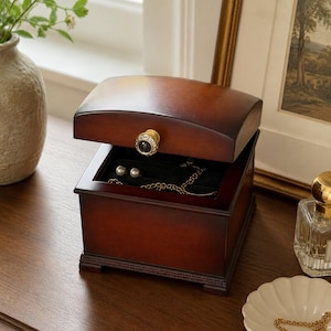 May include: A dark brown wooden jewelry box with a curved lid and a gold-toned knob featuring a central black stone surrounded by clear crystals. The box is partially open, revealing a black velvet interior containing a gold chain and pearl earrings.