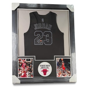 Op de afbeelding: Een ingelijste display met een zwart Michael Jordan Chicago Bulls shirt met de naam JORDAN en nummer 23 in grijs, een handtekening, twee actiefoto's van de speler en een rond Chicago Bulls logo-embleem in het midden.