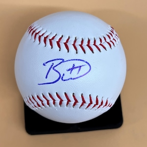 May include: A white baseball with red stitching and a blue autograph. The baseball is resting on a black stand against a tan background. The autograph appears to be the initials "B" and "H".