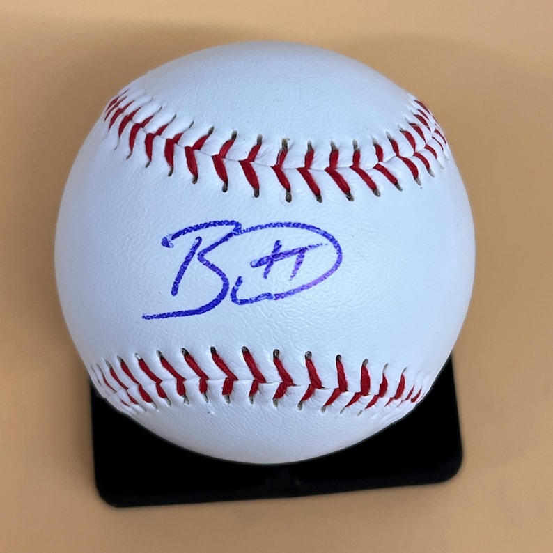 May include: A white baseball with red stitching and a blue autograph. The baseball is resting on a black stand against a tan background. The autograph appears to be the initials "B" and "H".