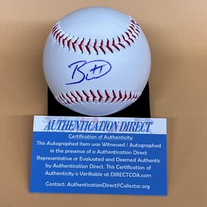 May include: A white baseball with red stitching and a blue signature. The ball is displayed on a black stand, with a blue and white "AUTHENTICATION DIRECT" certificate below it.