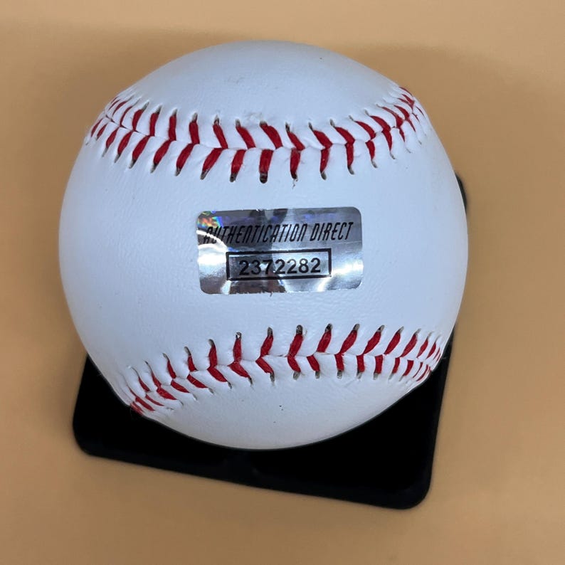 May include: A white baseball with red stitching, resting on a black stand. A silver sticker with the text "AUTHENTICATION DIRECT" and the number "2372282" is affixed to the ball.