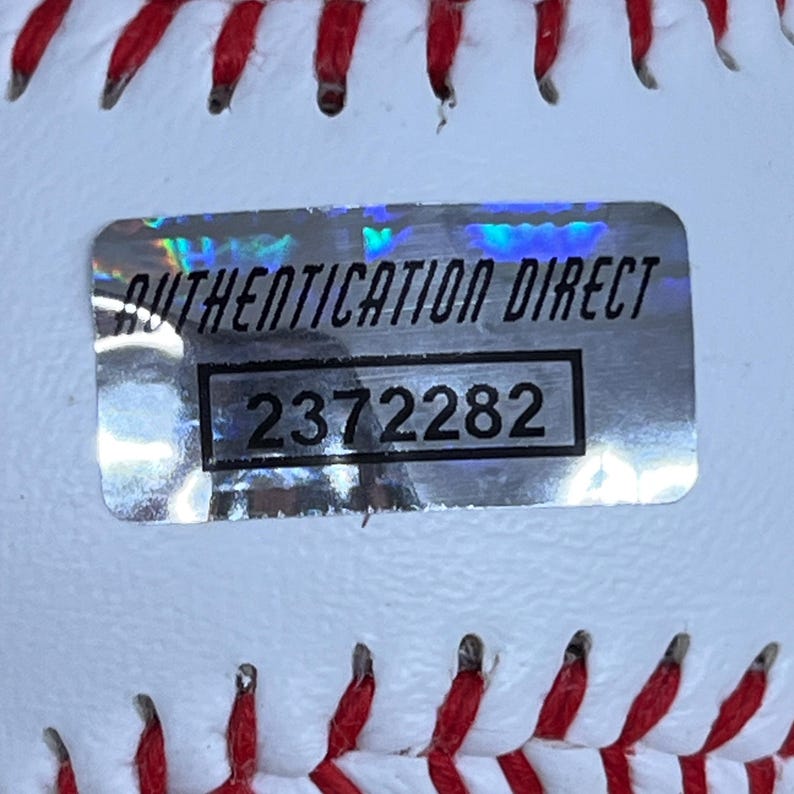 May include: Close-up of a white baseball with red stitching. A silver Authentication Direct sticker is affixed to the ball, displaying the text "AUTHENTICATION DIRECT" and the number "2372282".